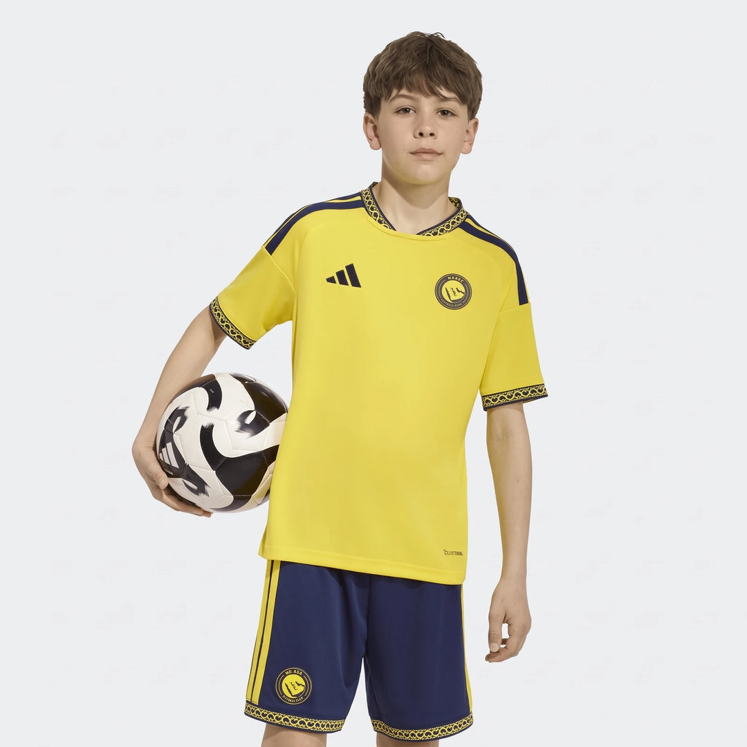Youth Al Nassr FC 26/27 home jersey in yellow and navy, worn by a child holding a soccer ball. Features Adidas logo and club