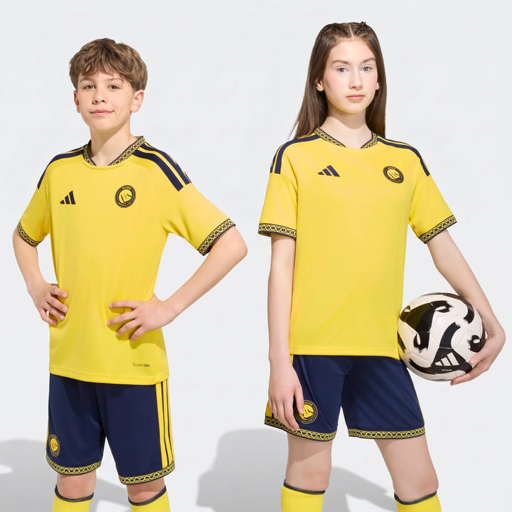 Two kids, a boy and a girl, wear the yellow and navy Al Nassr FC 26/27 Home Jersey soccer uniform with shorts and socks.