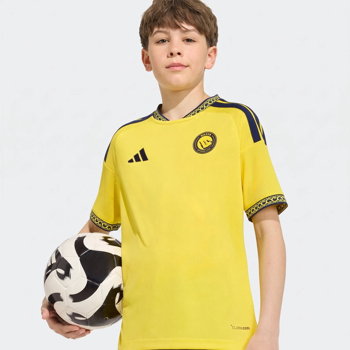 Young boy modeling an Al Nassr Fc 26/27 Home Jersey in yellow and navy, holding a soccer ball.