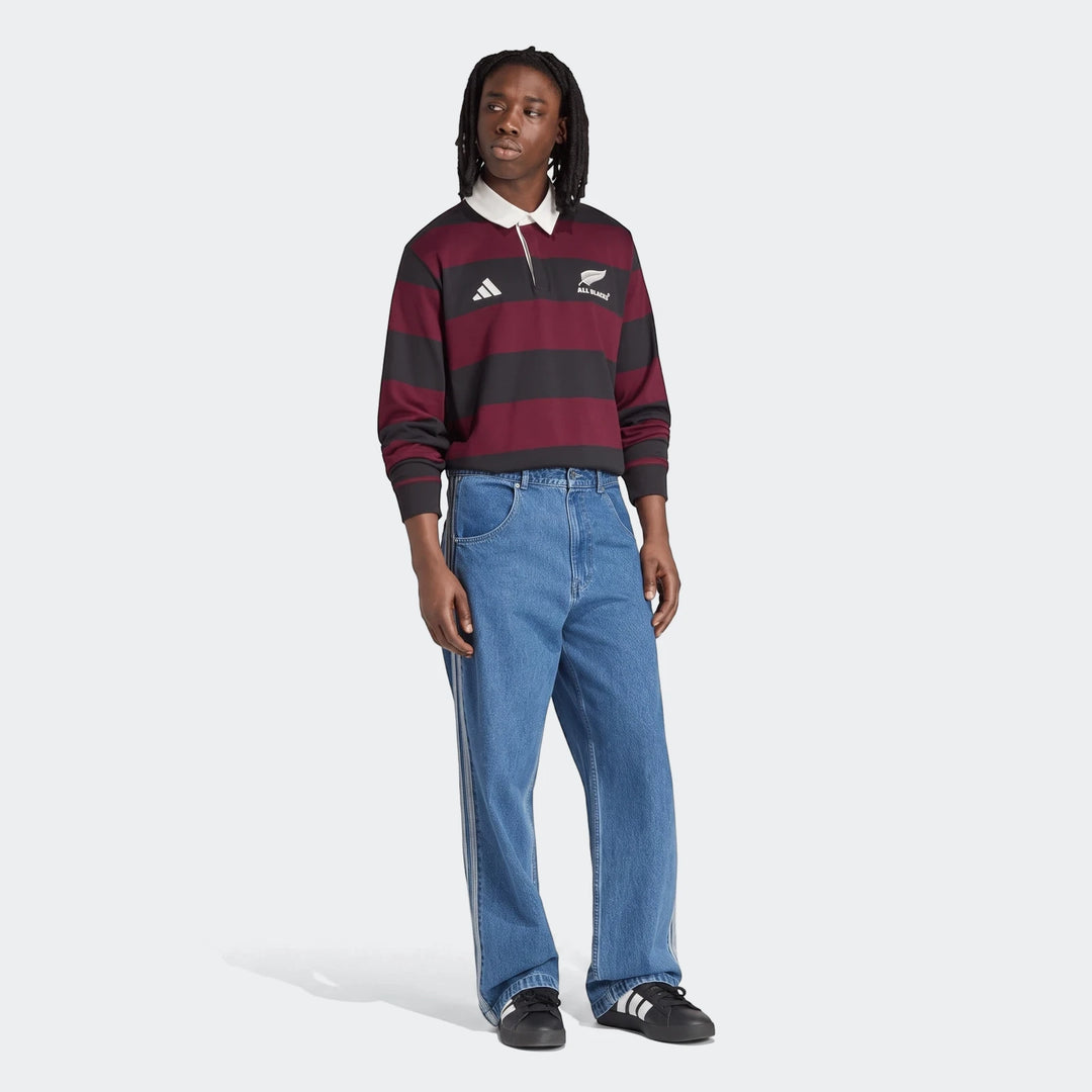 Male model wearing an Adidas All Blacks Heritage maroon and black striped long-sleeve rugby polo shirt, blue denim jeans,