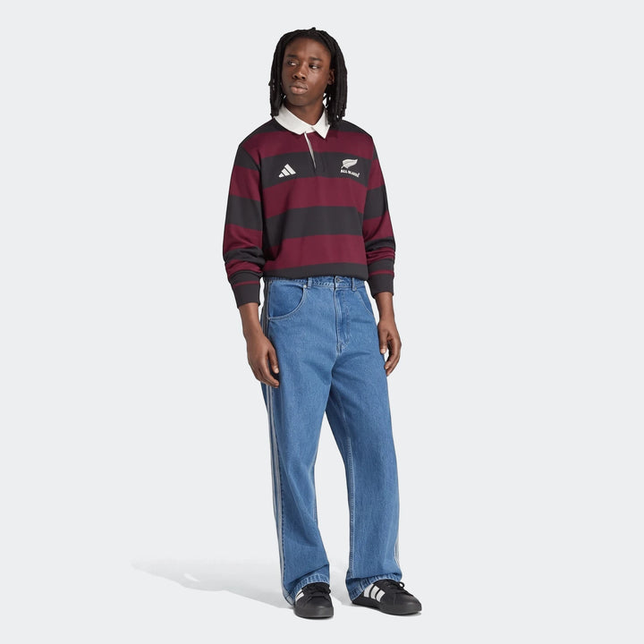 Male model wearing an Adidas All Blacks Heritage maroon and black striped long-sleeve rugby polo shirt, blue denim jeans,