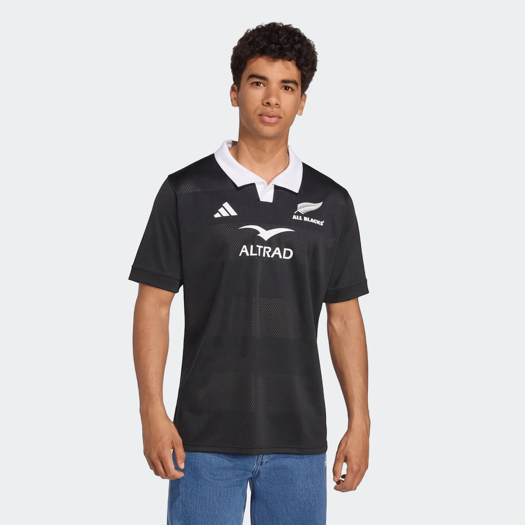 Man wearing a black Adidas All Blacks Home Jersey with white collar and sponsor logos, featuring short sleeves.