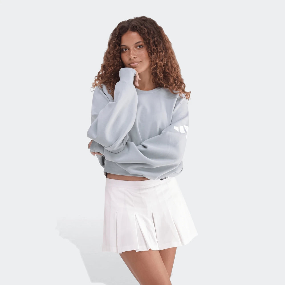 A female model wearing a light blue All Season Slim Sweatshirt and a white pleated mini skirt, posing with one hand on her