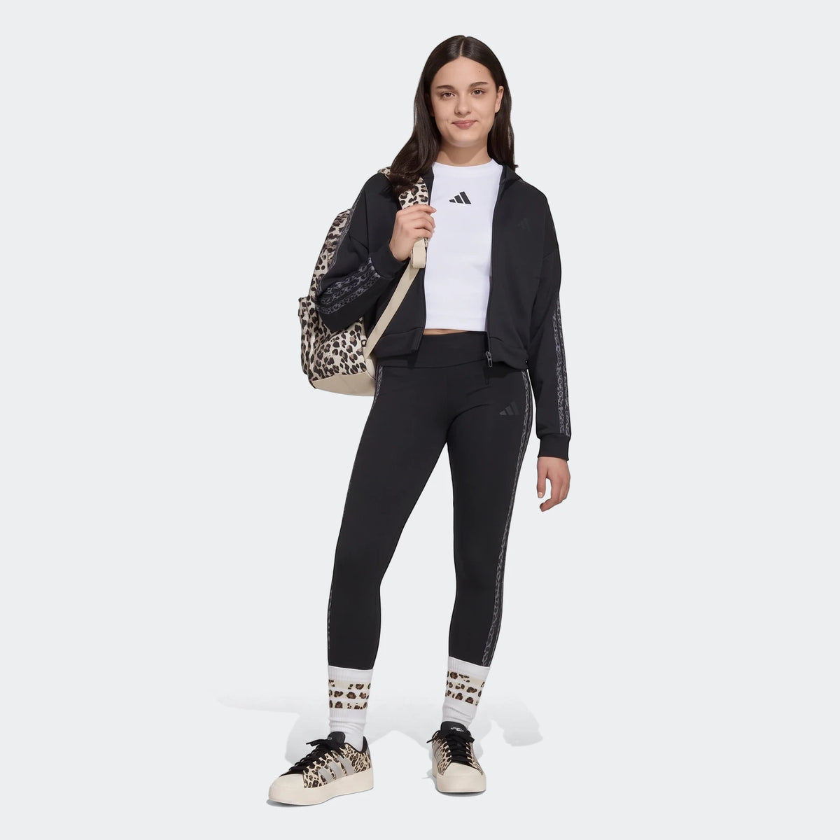 Young woman wearing black Adidas Animal Leggings, a black cropped hoodie, white t-shirt, and leopard print sneakers,