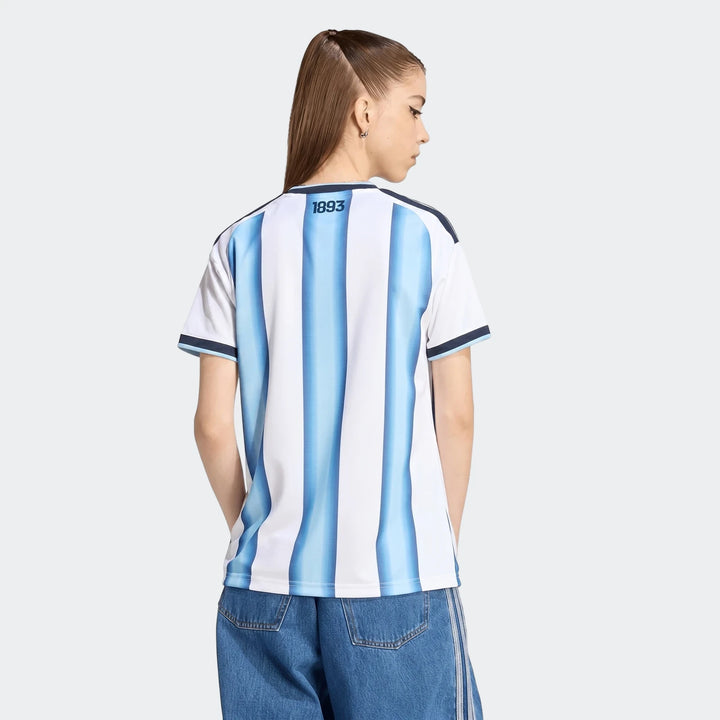 Argentina 26 Home Jersey worn by a female model, shown from the back with blue and white vertical stripes. Model wears blue
