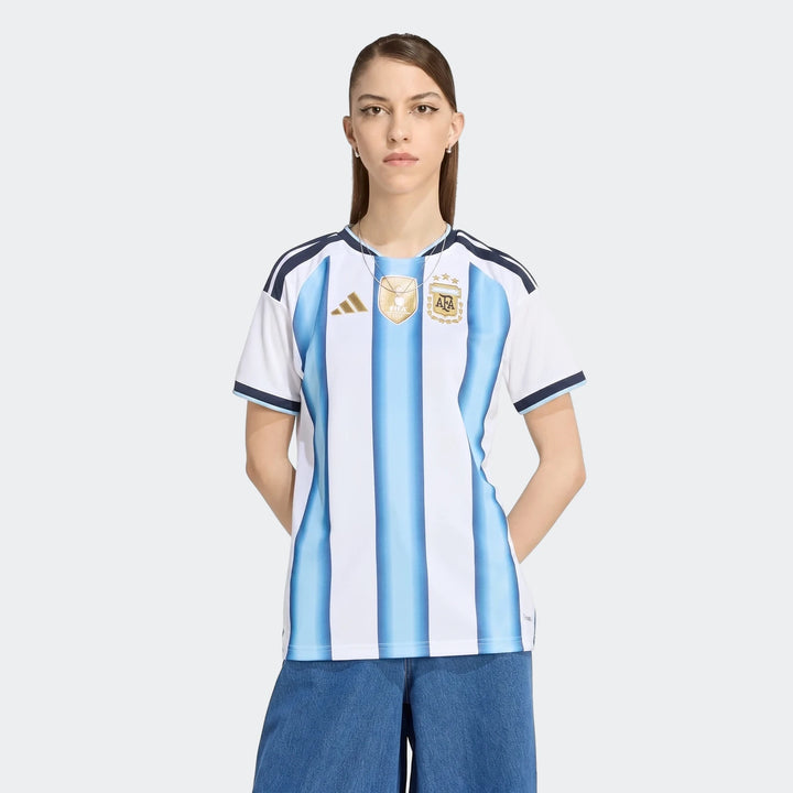 Female model wearing the Argentina 26 Home Jersey, featuring white and light blue stripes, with FIFA World Champions badge.