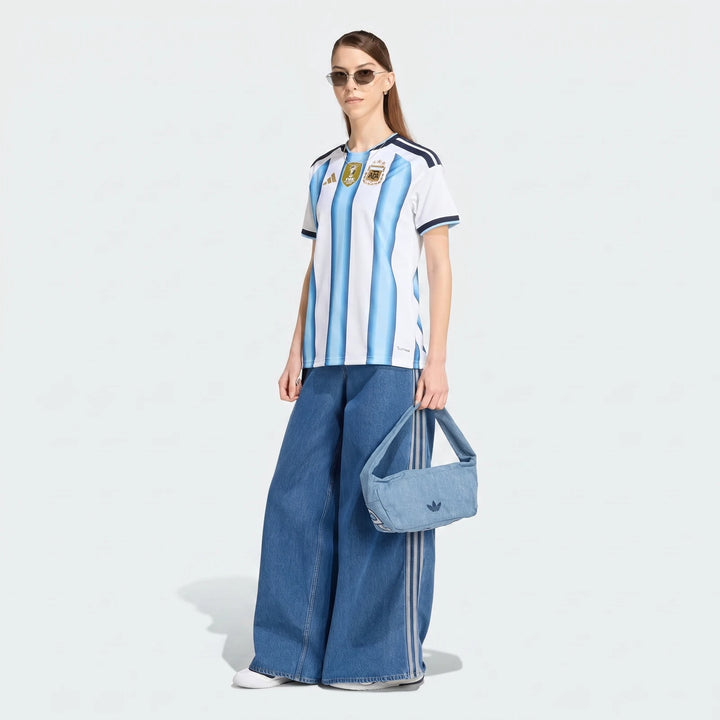 Model wearing the Argentina 26 Home Jersey, wide-leg denim pants, white sneakers, sunglasses, and a light blue denim