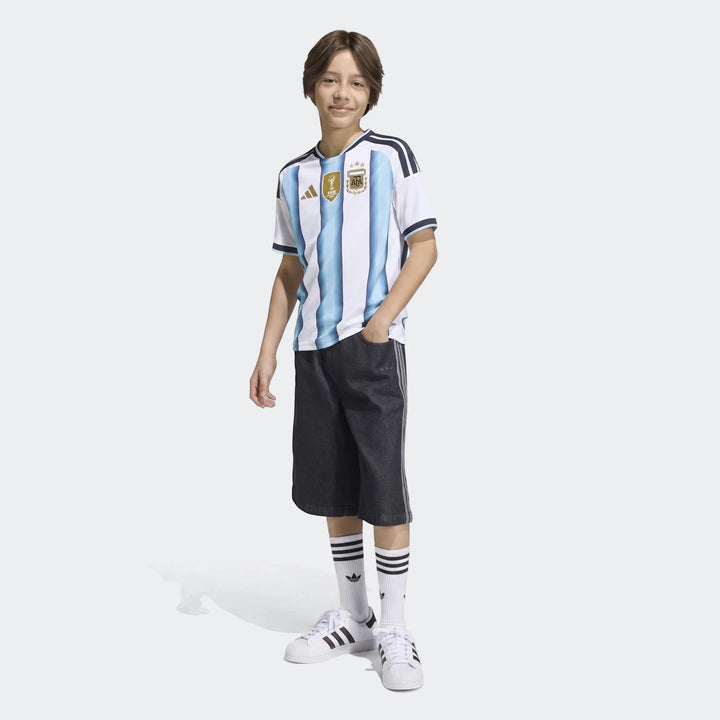 Young boy wearing a white and light blue striped Adidas Argentina 26 Home Kids Jersey, featuring a FIFA 2022 badge.