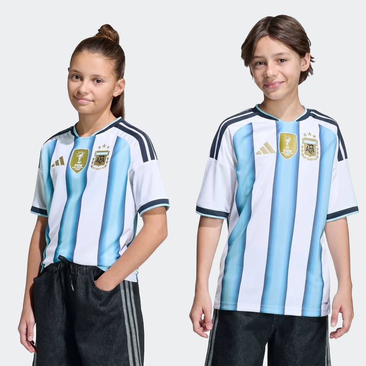 Two kids, a girl and a boy, model the Argentina 26 Home Kids Jersey, featuring white and light blue stripes with team logos.