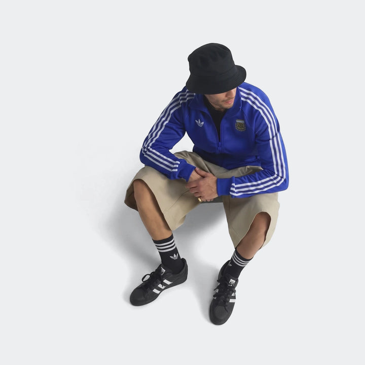 A man seated, wearing a blue Adidas Argentina Originals Track Top with white stripes, beige shorts, and black sneakers.