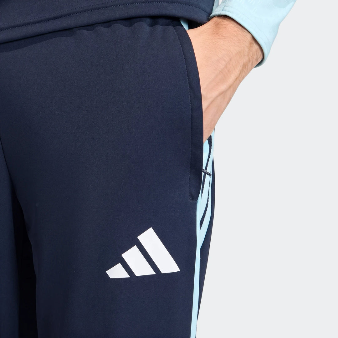 Close-up of dark blue Adidas Argentina training pants featuring light blue stripes, with a person's hand casually in the