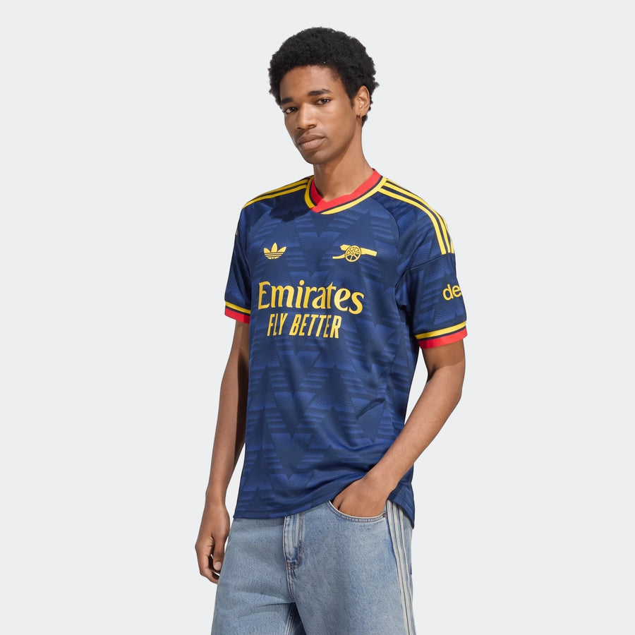 Male model wearing the dark blue Arsenal FC 26/27 Away Jersey, featuring yellow branding and red trim, posed waist-up.
