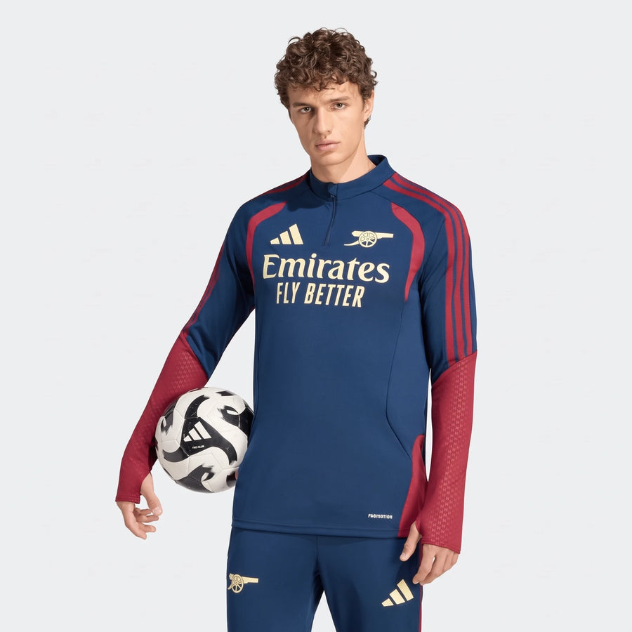 Male model wearing an Arsenal FC 26/27 Cup Tiro Training Top in navy blue, red, and gold, holding a soccer ball.