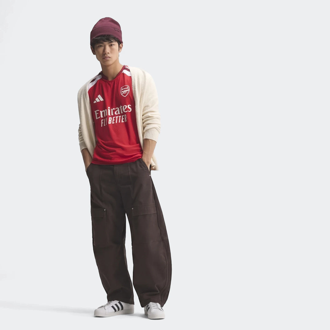 Male model wearing Arsenal FC 26/27 Home Authentic Jersey with a white cardigan, brown cargo pants, and white sneakers, full