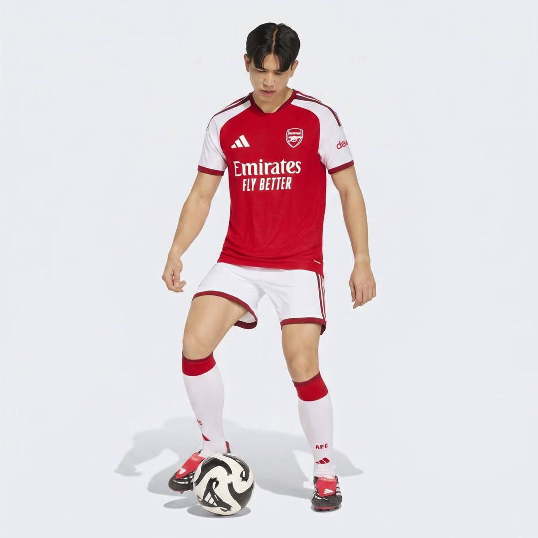 Male model wearing an Arsenal FC 26/27 Home Authentic Jersey, red and white, in a full body shot with a soccer ball.