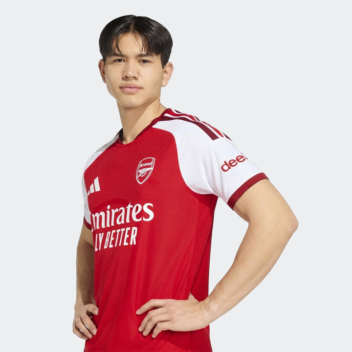 Young man wearing an Arsenal Fc 26/27 Home Authentic Jersey in red and white, posing with hands on hips.