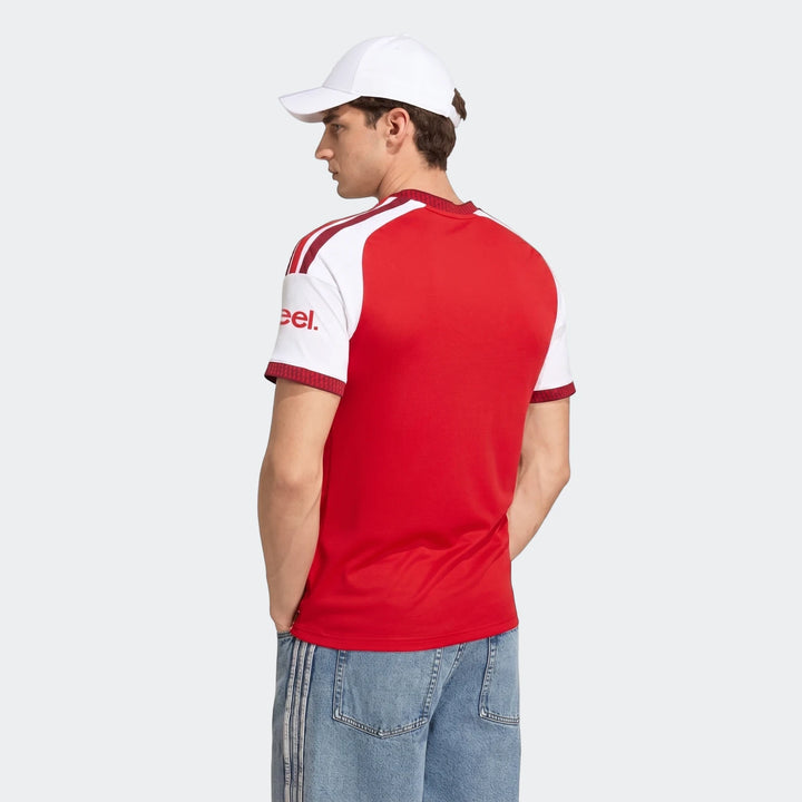 Back view of a male model wearing an Arsenal FC 26/27 home jersey in red and white with a white cap and blue jeans.