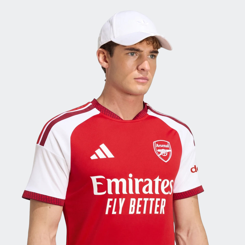 Male model wearing an Arsenal FC 26/27 Home Jersey in red and white, paired with a white baseball cap, against a neutral