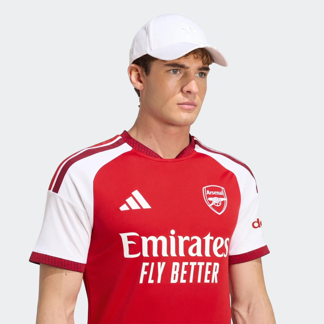 Male model wearing an Arsenal FC 26/27 Home Jersey in red and white, paired with a white baseball cap, against a neutral