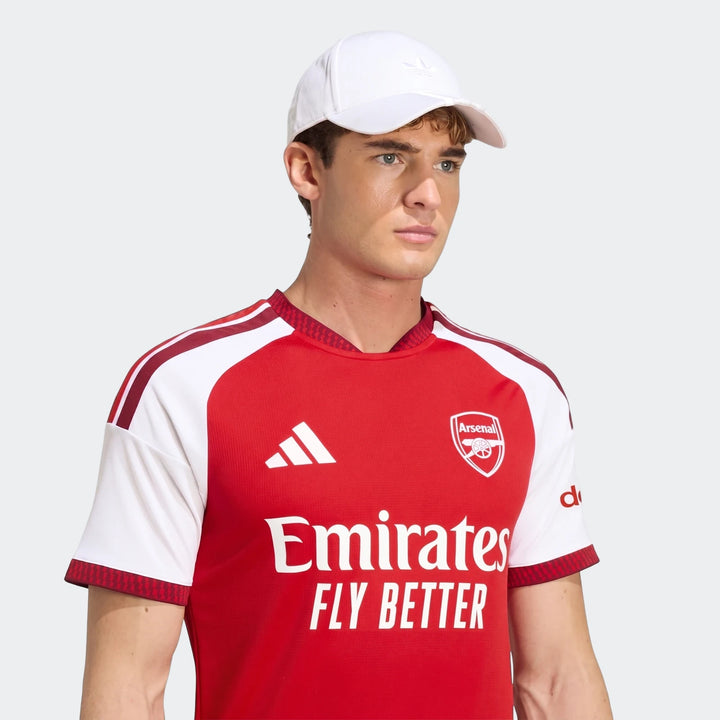 Male model wearing an Arsenal FC 26/27 Home Jersey in red and white, paired with a white baseball cap, against a neutral