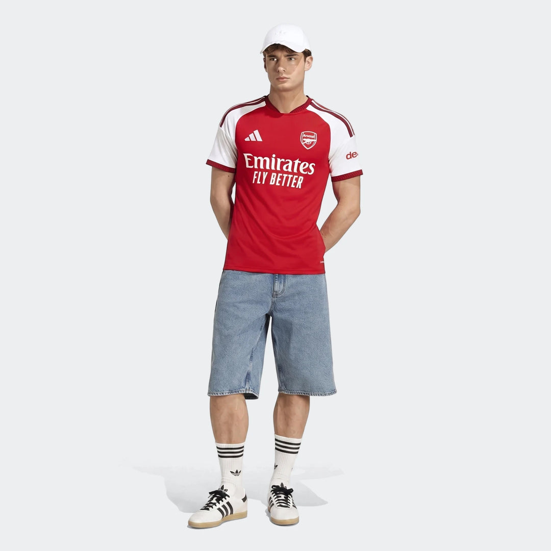 Male model wearing the new Arsenal FC 26/27 Home Jersey in red and white, paired with denim shorts, white socks, and