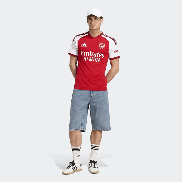 Male model wearing the new Arsenal FC 26/27 Home Jersey in red and white, paired with denim shorts, white socks, and