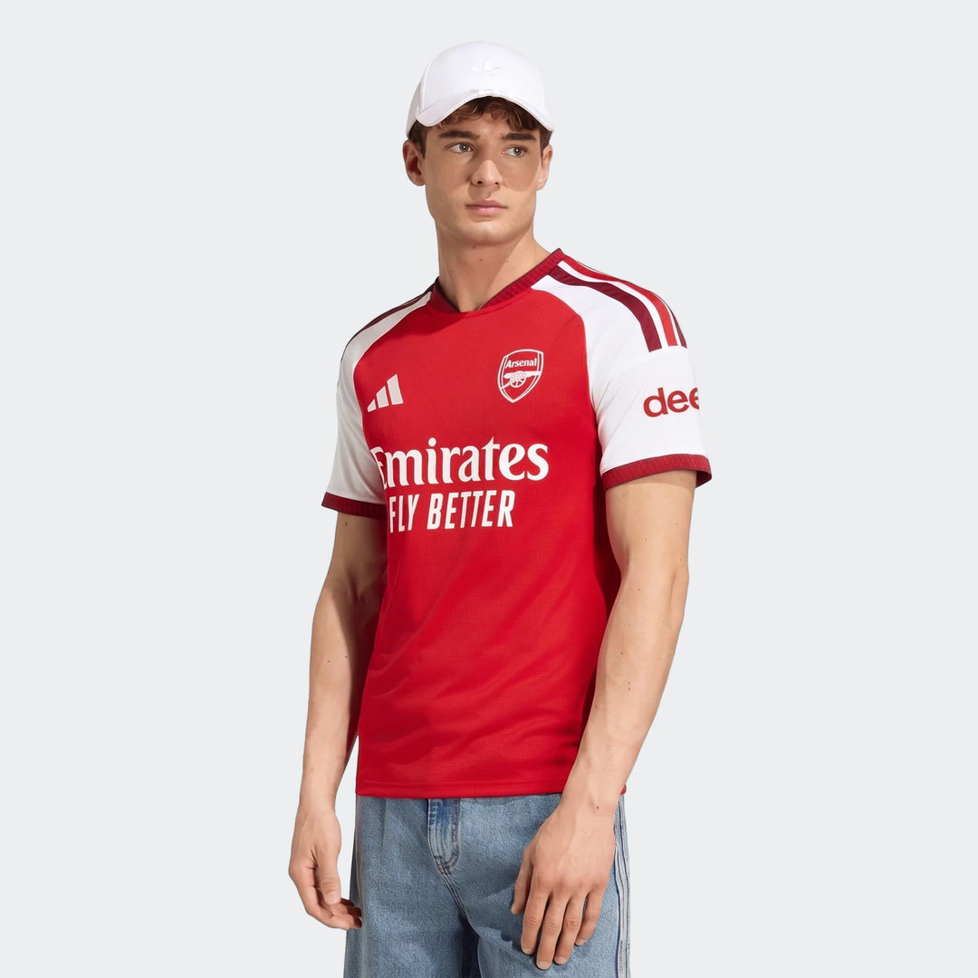 Young man modeling the red and white Arsenal FC 26/27 Home Jersey with blue jeans and a white cap.