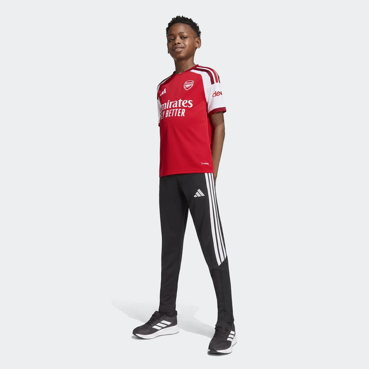 Young boy wearing a red Arsenal FC 26/27 Home Kids Jersey with black Adidas track pants and sneakers, full body view.