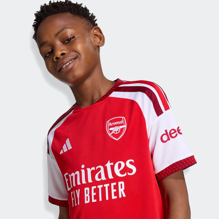 Young boy wearing the red and white Arsenal FC 26/27 Home Kids Jersey, smiling. Features the Arsenal crest and sponsor logos.