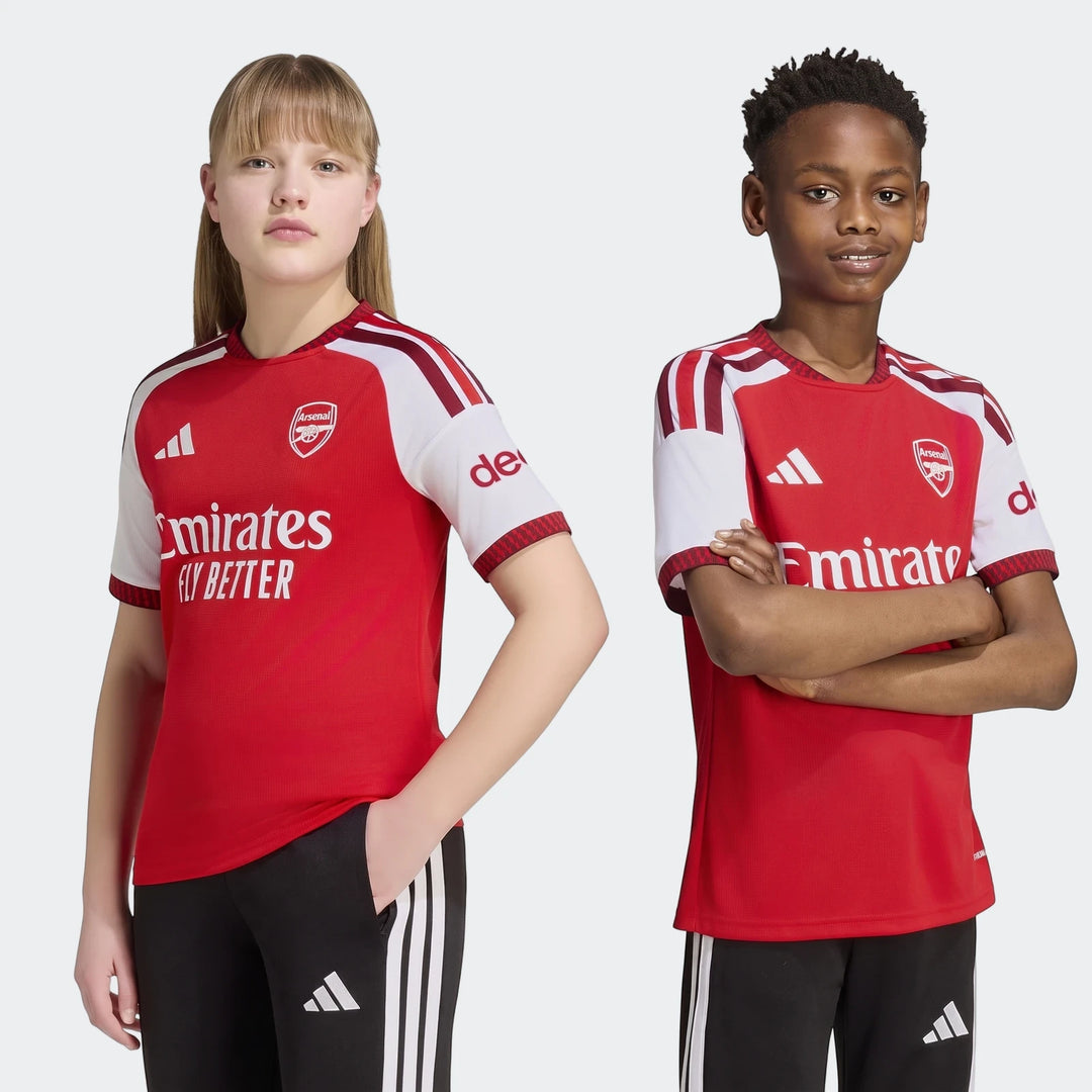 Two children, a boy and a girl, model the red and white Arsenal Fc 26/27 Home Kids Jersey and black track pants.