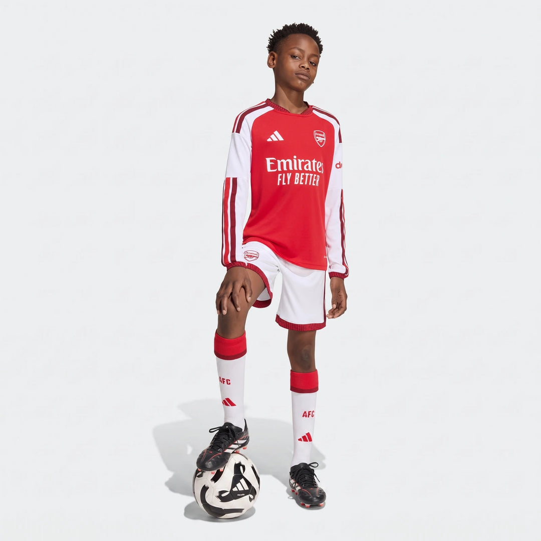 Young boy models Adidas Arsenal FC 26/27 home kit, featuring red and white long-sleeve jersey, white shorts, socks, and