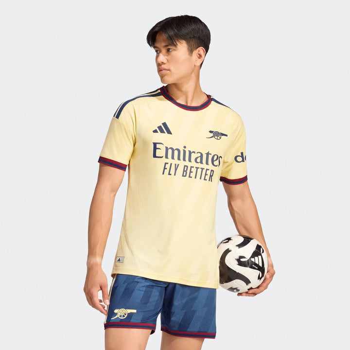 Male model wearing an Arsenal FC 26/27 Third Authentic Jersey in yellow, with blue shorts, holding a black and white soccer