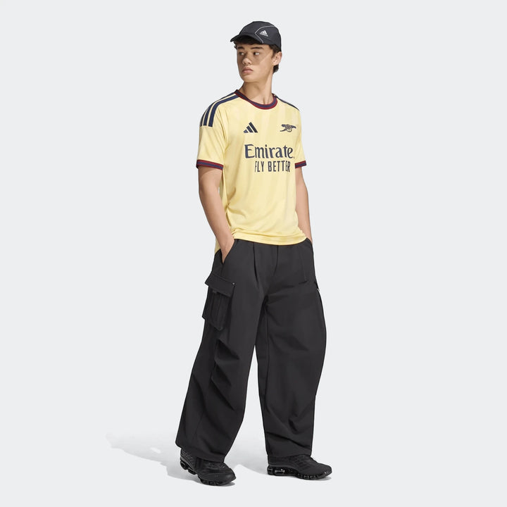 Male model wearing the yellow Adidas Arsenal FC 26/27 Third Jersey with black cargo pants and sneakers, full body shot.