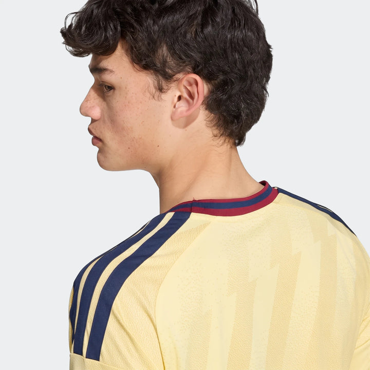Young male model in profile wearing a light yellow Arsenal Fc 26/27 Third Jersey with blue shoulder stripes.