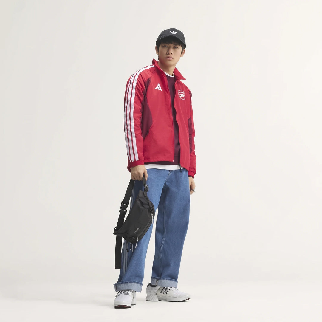 East Asian male model wearing a red Arsenal FC Home Anthem Jacket, blue jeans, and a black cap, holding a fanny pack.