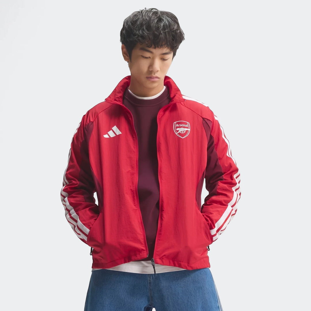 Young man in a red and maroon Adidas Arsenal FC Home Anthem Jacket with white stripes and team crest, paired with blue jeans.