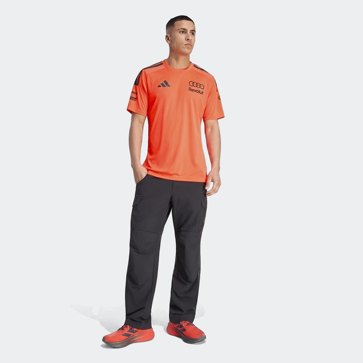 Man wearing an orange Audi Revolut F1 Team Setup T-Shirt, dark gray cargo pants, and orange sneakers, standing full-body.