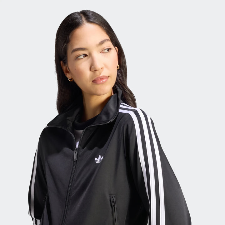 A woman models a black Adidas Firebird Classic Track Jacket with iconic white three-stripes on the sleeves and a Trefoil