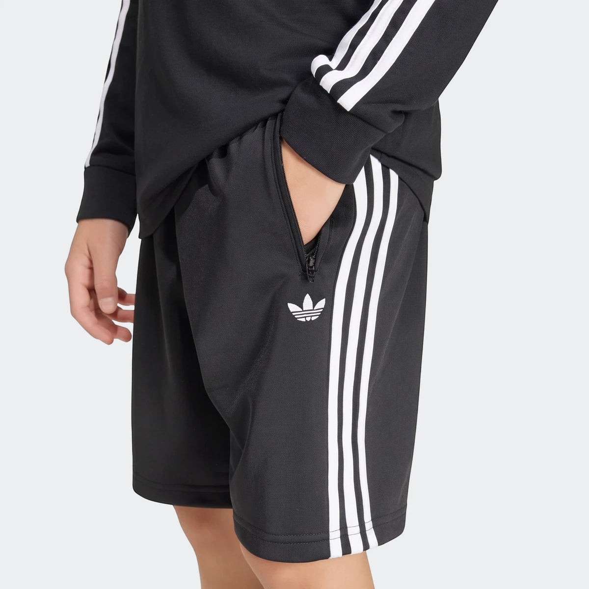 Close-up of black Adidas Firebird shorts with white three stripes, worn by a person, featuring a zippered pocket and Trefoil