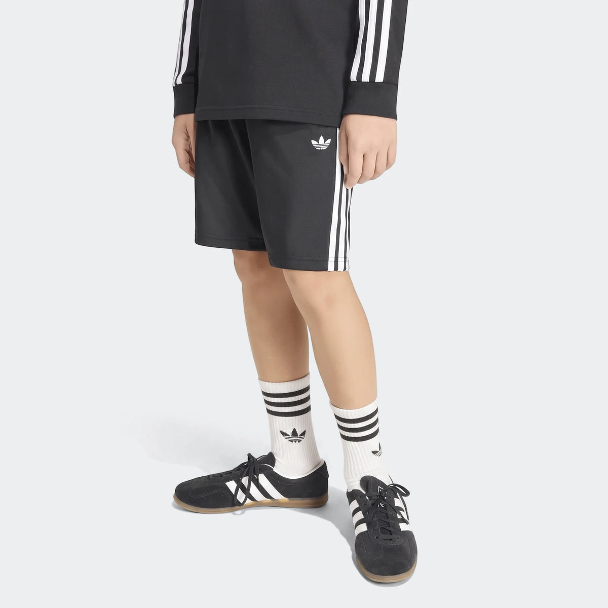 Black Adidas Firebird shorts with white stripes, worn by a model, paired with a black long-sleeve shirt, white socks, and