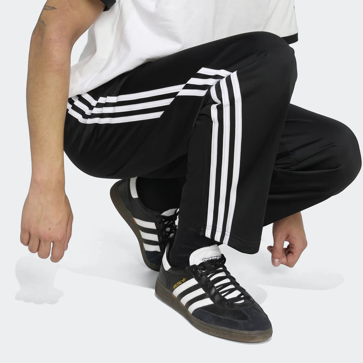 Black Adidas Firebird track pants with white three-stripe detailing, worn by a person crouching and paired with black and