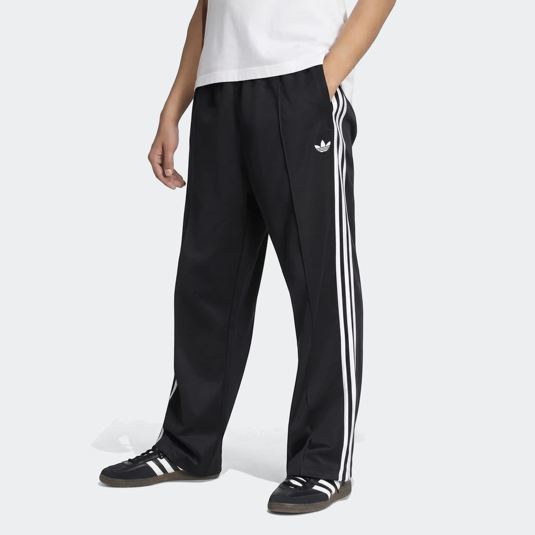 Black Adidas Firebird Track Pants with white three-stripe design worn by a model, showcasing a classic athletic style.