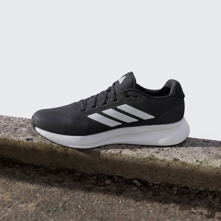 Black Adidas running shoe with white three-stripe logo and white sole, displayed on a concrete surface outdoors.