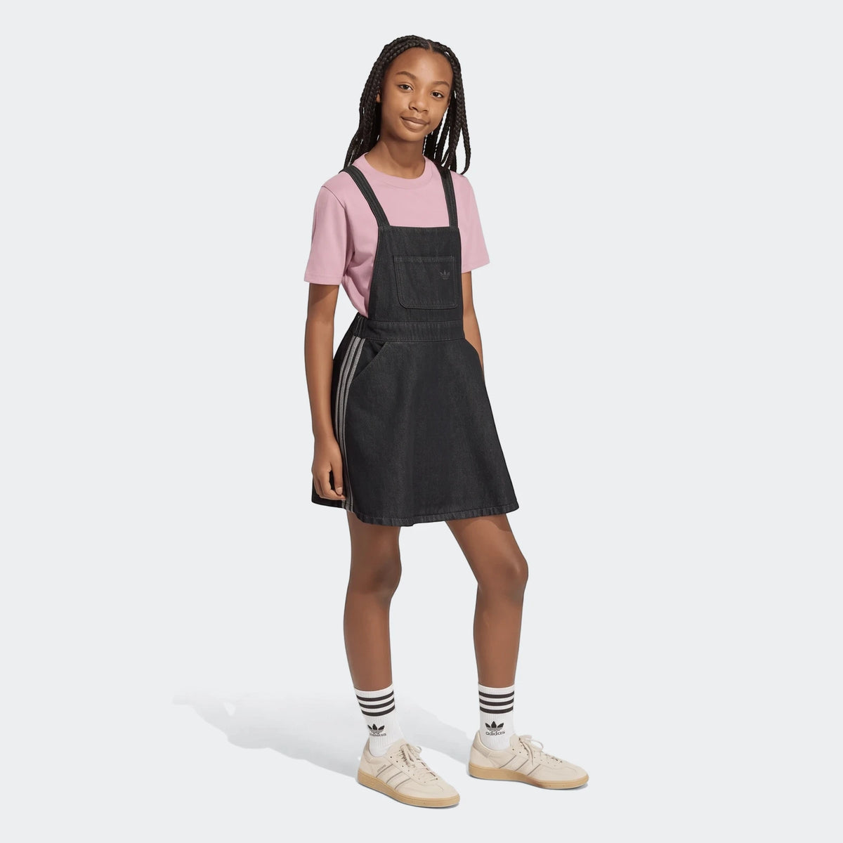Young person wearing a black denim dungarees dress over a pink t-shirt with white striped socks and beige sneakers, full