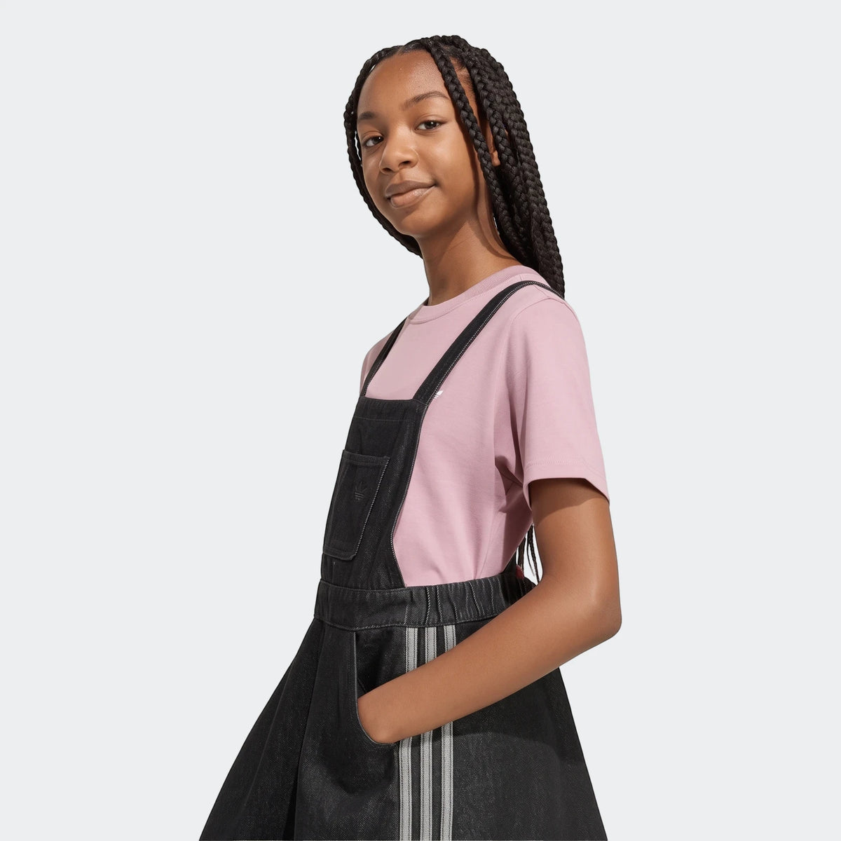 Young woman wearing a black denim dungarees dress over a pink t-shirt, posing with her hand in the pocket.