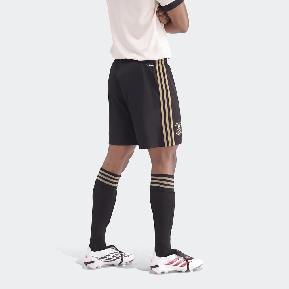 Black and gold soccer shorts with three stripes and a skull logo, worn by a model with black socks and white cleats.