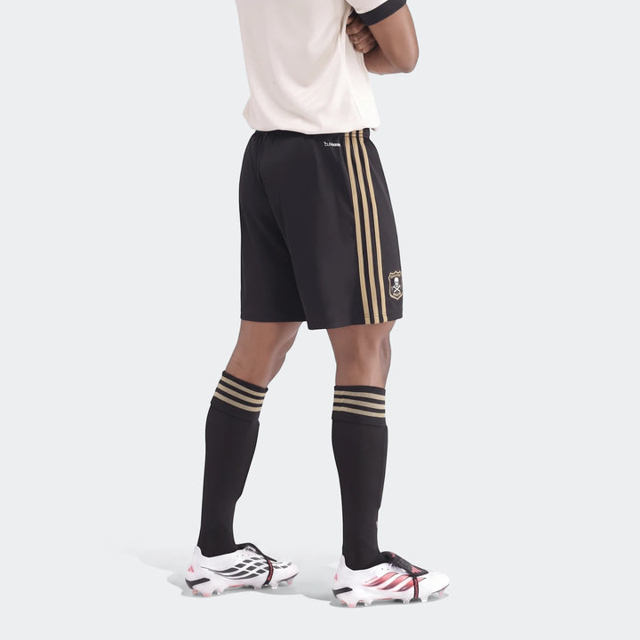 Black and gold soccer shorts with three stripes and a skull logo, worn by a model with black socks and white cleats.