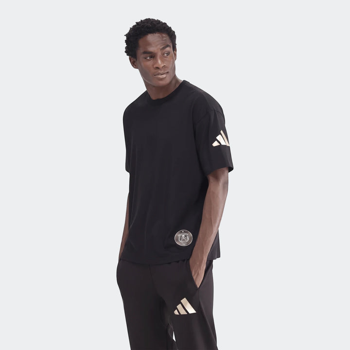 A male model wears a black Sportswear Open Hem Single Jersey T-Shirt with a golden Adidas logo and black joggers, casually