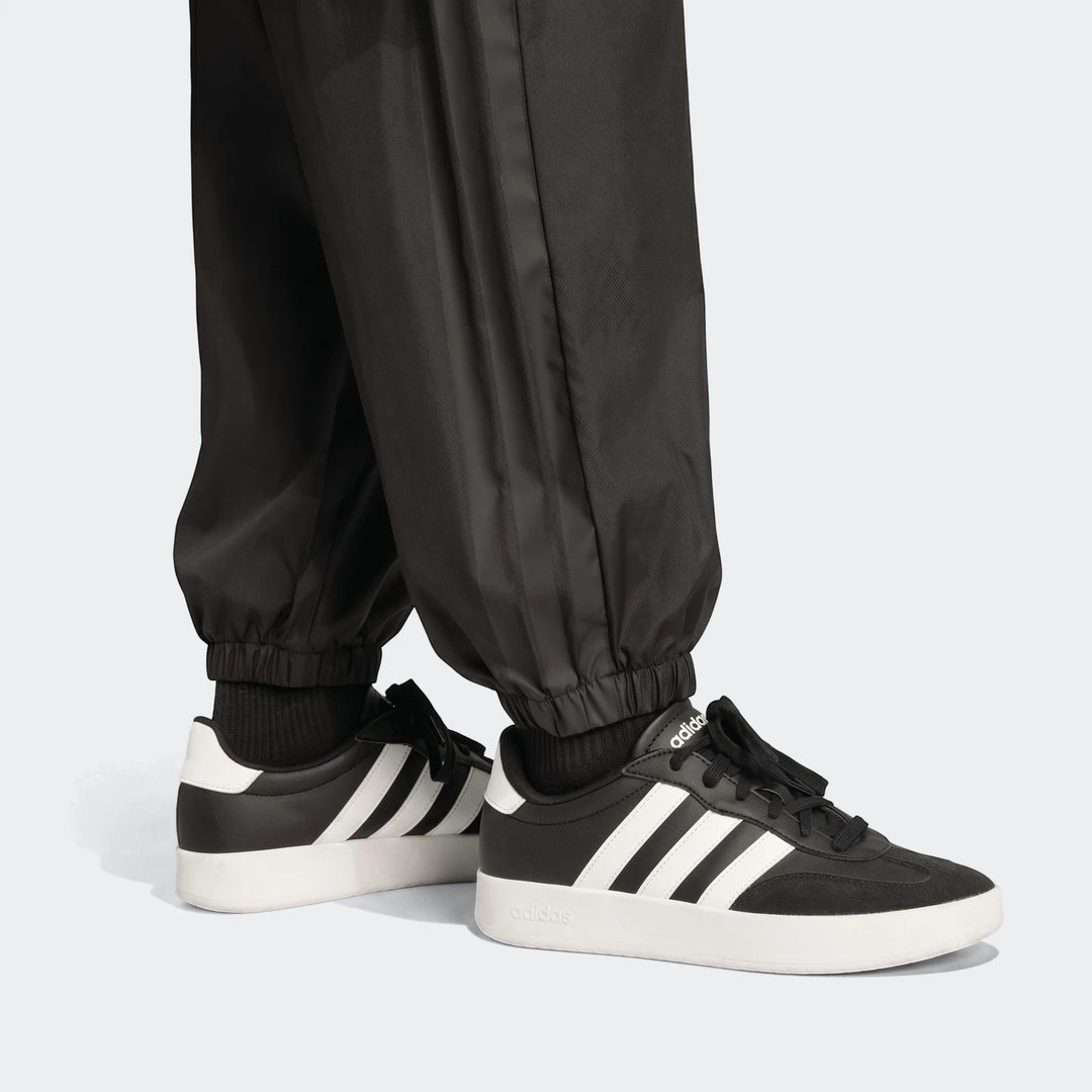Close-up of a person wearing black Utility Woven Pants with white three-stripes and black and white Adidas sneakers.