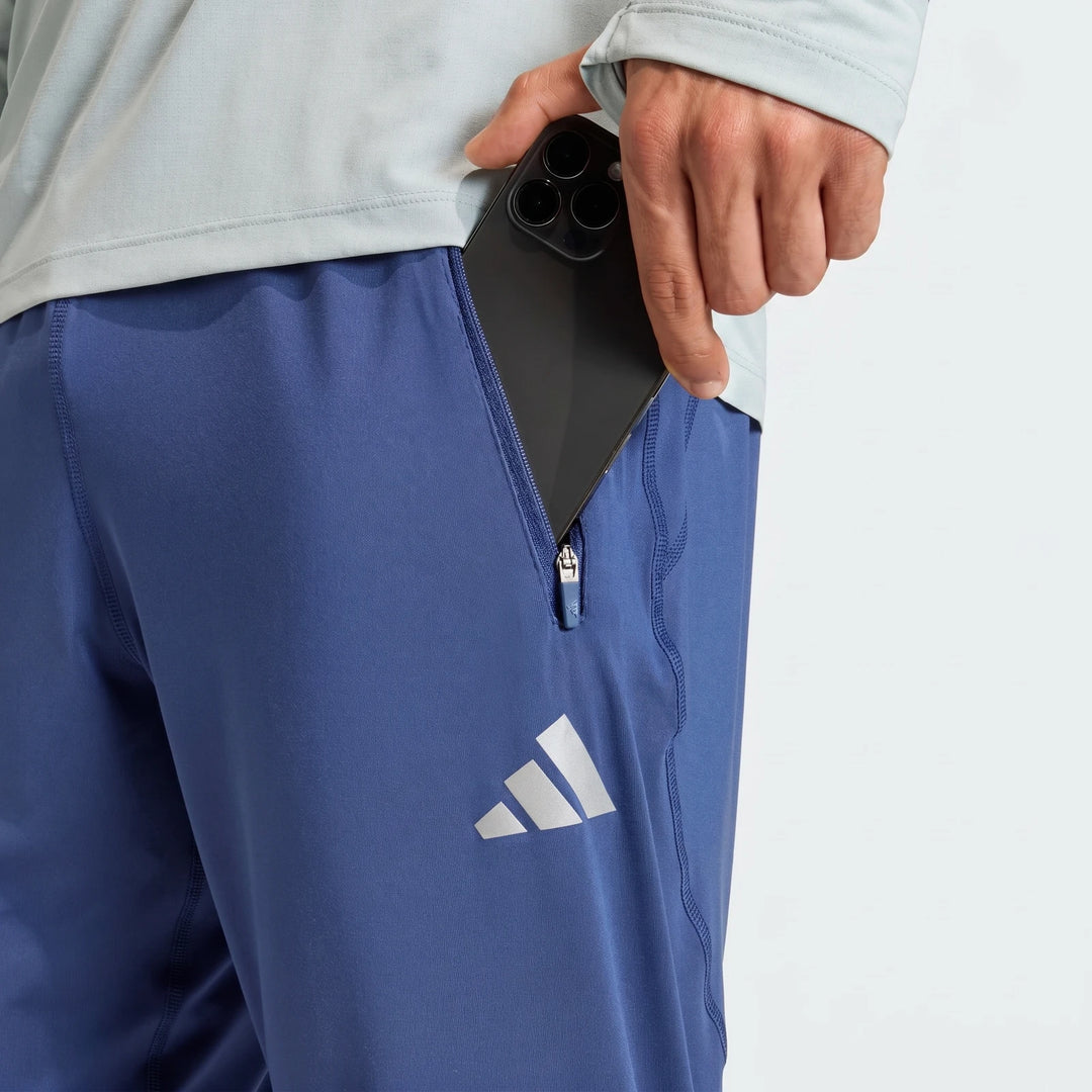 A close-up shot of a man wearing blue Adidas Adi365 Track Pants, placing a black smartphone into a zippered side pocket.
